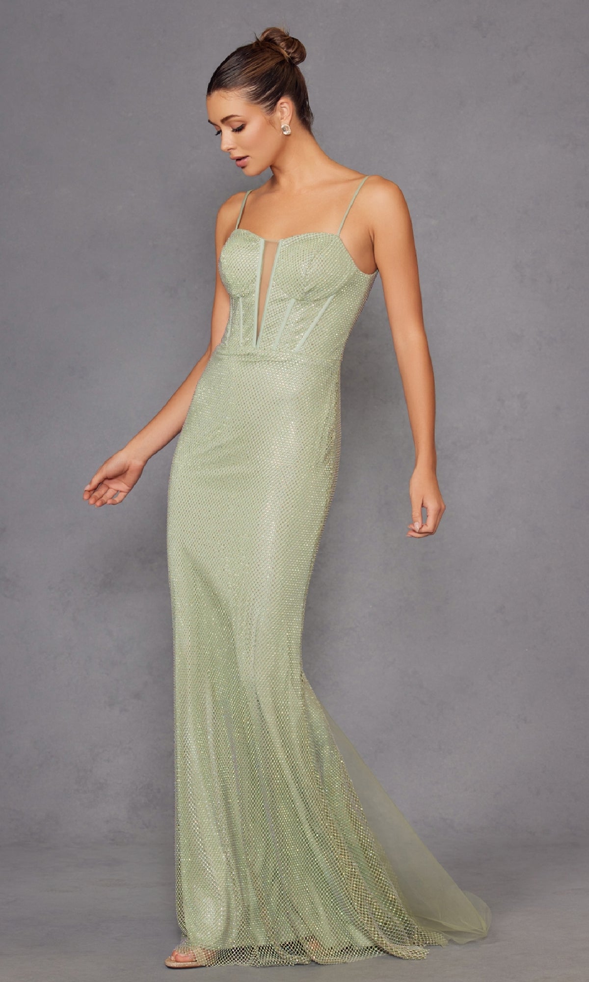 A woman models the Long Corset Beaded Prom Dress: Juliet JT2470S, a light green floor-length gown with thin straps and a sheer neckline panel, standing against a plain gray background.